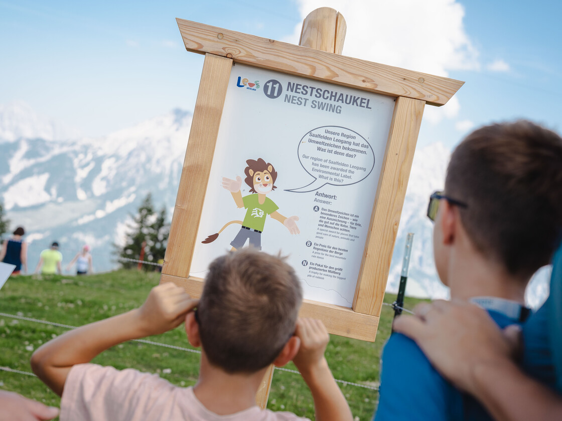 Kinder lesen die Rätselstation „Nestschaukel“ bei Leos Rätseljagd am Berg der Sinne in Leogang – Familienerlebnis mit Panorama auf die Loeganger Steinberge  | © Michael Geißler