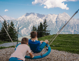 Zwei Kinder auf einer großen Nestschaukel am Berg der Sinne in Leogang mit Blick auf die Leoganger Steinberge | ©  Michael Geißler