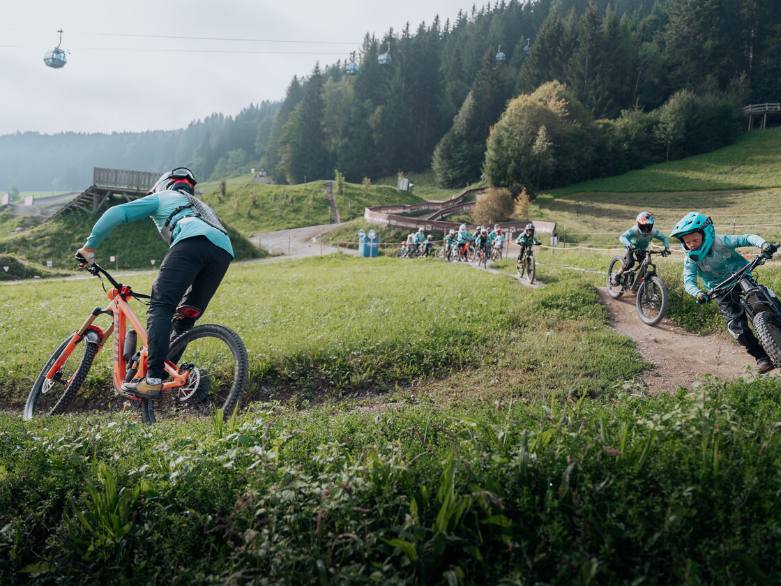 Bikeclub Leogang – Nachwuchs auf dem Trail | © Yvonne Hörl Junge Mountainbiker:innen fahren in einer Gruppe auf einem Flow-Trail im Bikepark Leogang, begleitet von einem Coach, mit Seilbahn und grüner Landschaft im Hintergrund. | © Yvonne Hörl
