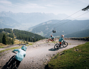 Steinberg Line by FOX Abfahrt im Bikepark Leogang | © Yvonne Hörl Junge Downhillerinnen fahren eine kurvige Abfahrt auf der Steinberg Line by FOX im Bikepark Leogang, mit beeindruckendem Alpenpanorama und Seilbahn im Hintergrund. | © Yvonne Hörl