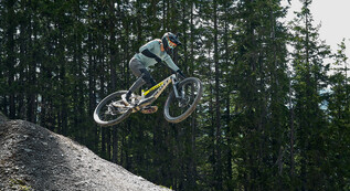 Mountainbikerin mit Vollvisierhelm und Protektoren springt mit ihrem Downhill-Bike über einen Kicker im Bikepark, umgeben von dichtem Wald. | © Karin Pasterer