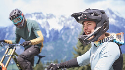 Gruppe von Mountainbiker:innen mit Vollvisierhelmen und Schutzkleidung posiert lachend vor einem Saalfelden-Leogang-Schild mit alpinem Panorama. | © Karin Pasterer