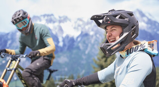 Gruppe von Mountainbiker:innen mit Vollvisierhelmen und Schutzkleidung posiert lachend vor einem Saalfelden-Leogang-Schild mit alpinem Panorama. | © Karin Pasterer