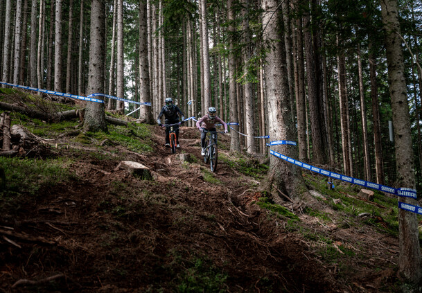 Zwei Biker:innen auf wurzeligem Steilstück in "Back to the Roots". | © Yvonne Hörl Zwei Mountainbiker:innen fahren mit vollem Fokus ein steiles, wurzeliges Waldstück entlang.  | © Yvonne Hörl