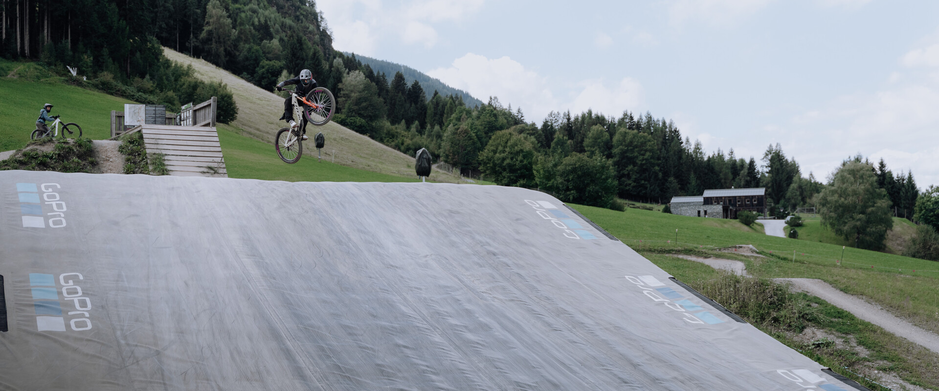 Ein Biker schiebt sein Rad über den Schotterweg in Richtung Bagjump-Rampe im Übungsareal bei der Talstation Leogang. Rechts ist das große GoPro-Luftkissen für sichere Landungen zu sehen. | © Yvonne Hörl