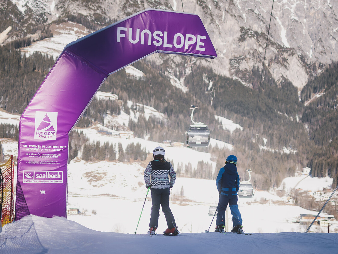 Zwei Kinder auf Skiern stehen am Start einer Funslope mit einem großen lila Torbogen, der den Eingang markiert. Im Hintergrund ist eine winterliche Berglandschaft mit Gondeln und Skipisten zu sehen. | © Stephanie Oberhauser