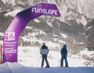 Zwei Kinder auf Skiern stehen am Start einer Funslope mit einem großen lila Torbogen, der den Eingang markiert. Im Hintergrund ist eine winterliche Berglandschaft mit Gondeln und Skipisten zu sehen. | © Stephanie Oberhauser