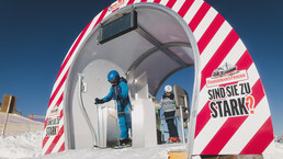 Zwei Skifahrer stehen am Start der Fisherman's Friend Skimovie Strecke und bereiten sich auf die Abfahrt vor. Der Eingang ist als futuristischer Tunnel mit einer auffälligen Fisherman’s Friend Werbung gestaltet. Die klare Berglandschaft und der blaue Himmel schaffen eine beeindruckende Kulisse. | © Stefanie Oberhauser