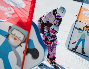 Erwachsene Person begleitet ein Kind durch einen Slalomparcours mit bunten Hindernisfiguren im Schnee. | © Michael Geißler
