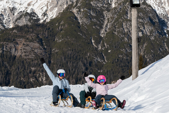 Familie beim Rodeln am Asitz in Leogang mit Blick auf das verschneite Steinerne Meer – Winterurlaub in Saalfelden Leogang | © cb photography / Christoph Bayer