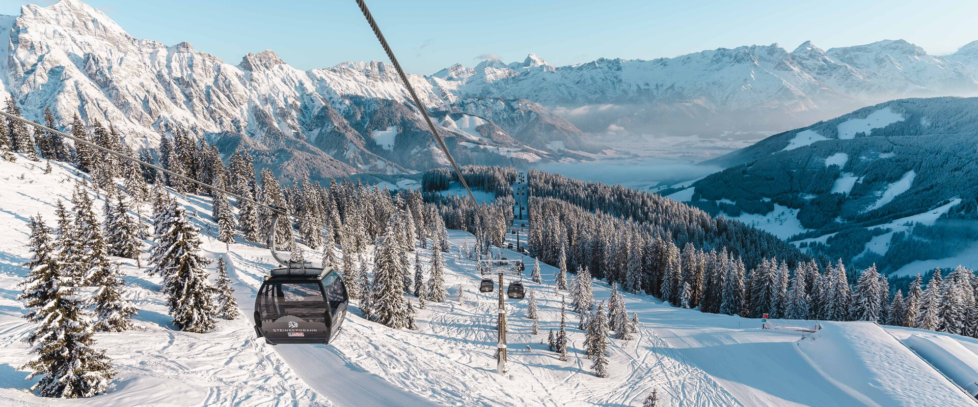 Dunkelgraue Steinberg Kabinenbahnen schweben über eine verschneite Skipiste am Asitz in Leogang. Dicht mit Schnee bedeckte Tannenwälder erstrecken sich entlang der Berghänge. Im Hintergrund ragen die mächtigen, weiß verschneiten Leoganger Steinberge und weitere Gipfel der Alpen in den hellblauen Himmel. Sonnenlicht und lange Schatten prägen die winterliche Landschaft. | © Michael Geißler